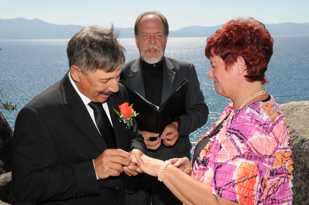 Groom places the wedding band on the finger of the bride while the minister watches