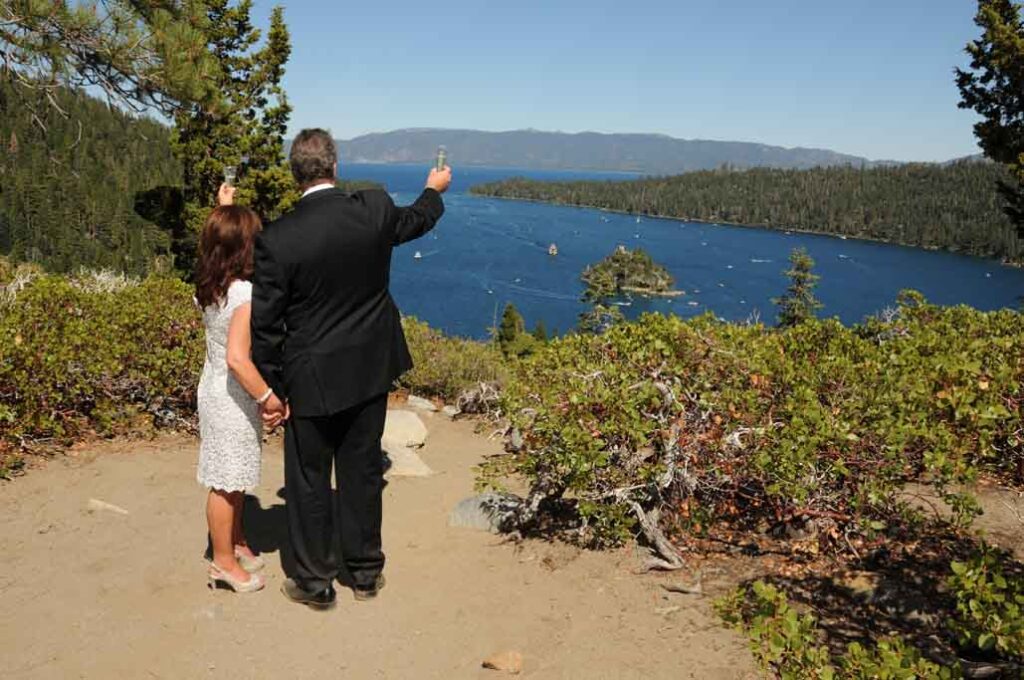 The married couple extend their arms while holding champagne glasses towards the lake